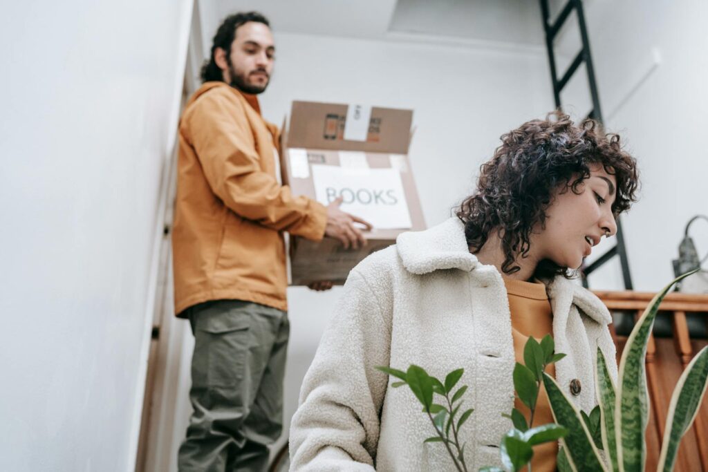 A couple carrying boxes and plants while moving; woman enjoys greenery.