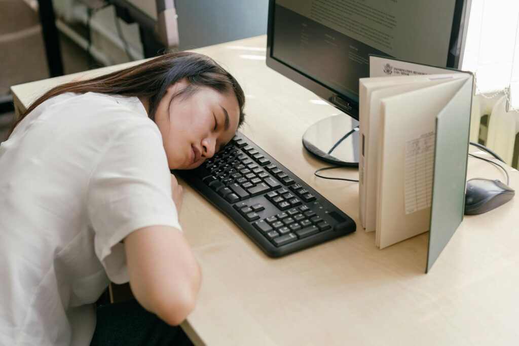 Young woman student asleep on a computer keyboard at her desk, surrounded by books.