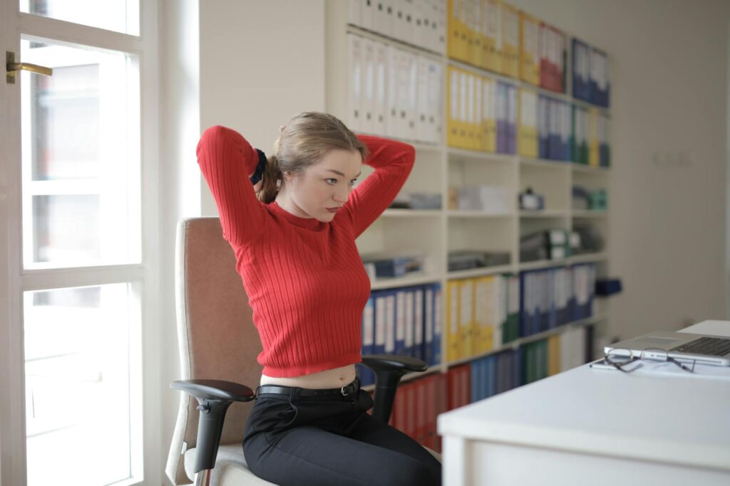 A young woman in a red sweater stretches at her desk in a modern office space.
