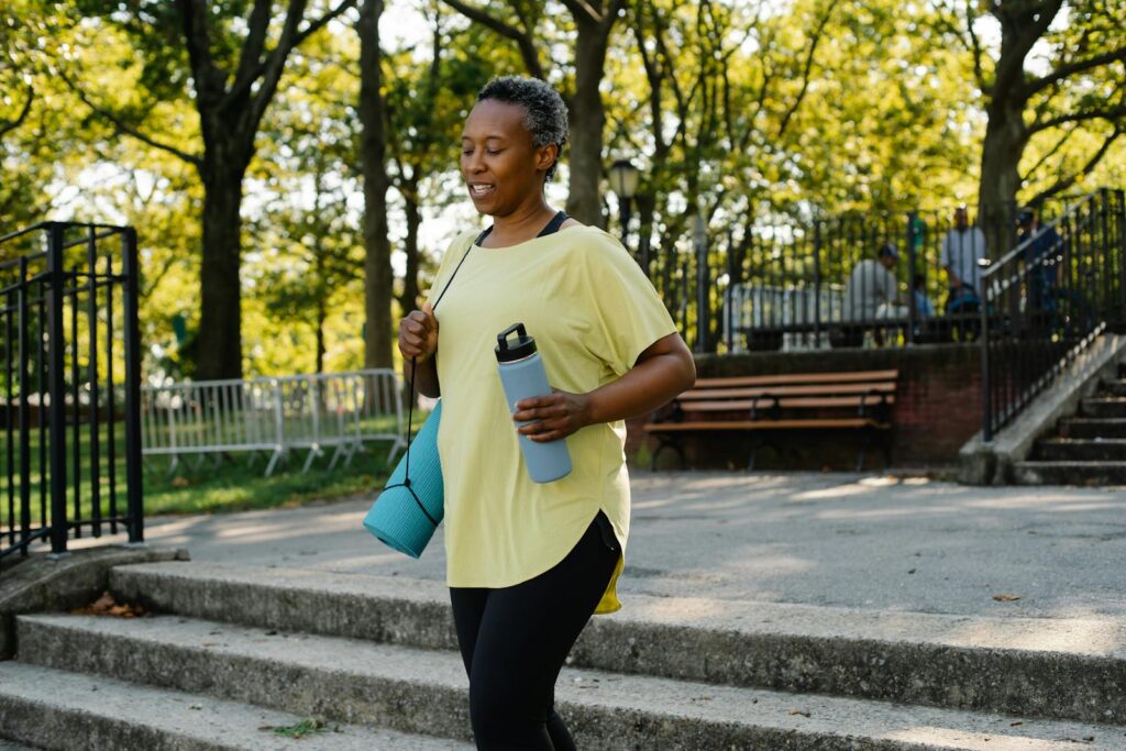Woman walking outdoors with a water bottle and yoga mat on a sunny day in a public park.