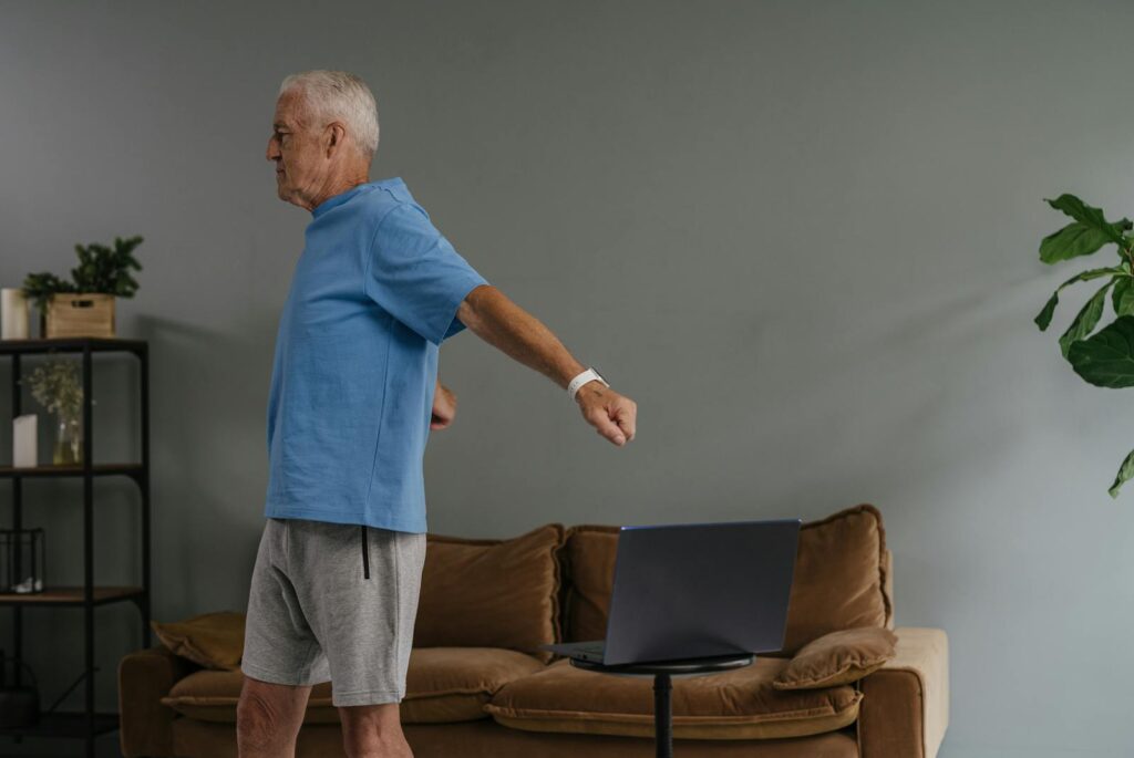 Elderly man stretching indoors while watching laptop, promoting active lifestyle.
