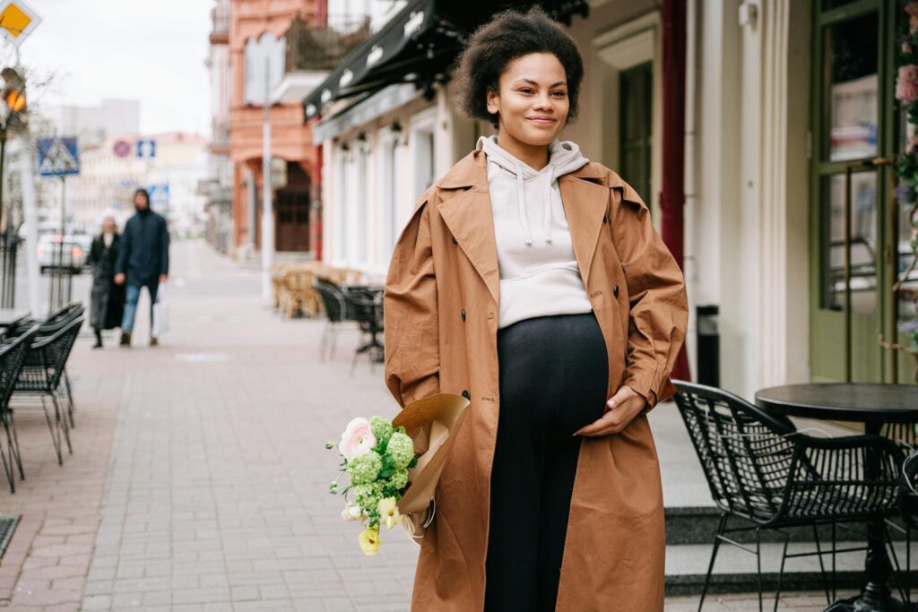 Smiling pregnant woman with bouquet walking in city street, wearing trench coat.